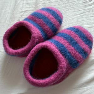 May include: A pair of cozy, striped slippers. The slippers are primarily magenta with horizontal blue stripes. The interior is a deep red. The slippers are made of a soft, textured material, perfect for indoor wear.
