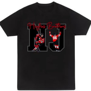 May include: Black t-shirt with a white and gray "NJ" graphic. The graphic features two hockey players in red and black jerseys with the text "Hughes Brothers" above.