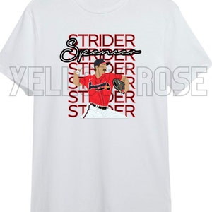 Peut inclure: T-shirt blanc avec un graphisme d'un joueur de baseball en uniforme rouge et marine. Le t-shirt porte le nom "Strider Spencer" dans une police stylisée. Le joueur est en position de lancer.