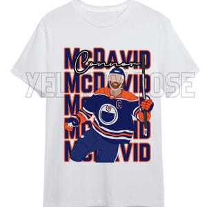 May include: White t-shirt featuring a graphic of a hockey player wearing a blue and orange jersey with the text "McDavid" repeated in a stacked pattern.