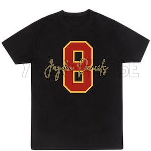 Puede incluir: Camiseta negra con un gran número 8 rojo con contorno dorado. El nombre "Jayden Daniels" está escrito en cursiva debajo del número. La camiseta es de estilo cuello redondo clásico.