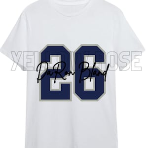 May include: White t-shirt with a navy blue and silver number 26 on the front. The text "Da Ron Bland" is written in black below the number.