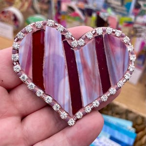 May include: A heart-shaped decorative piece with a mosaic design. The heart features vertical stripes of burgundy and lavender glass, framed by a pink border. The border is embellished with small, sparkling rhinestones.