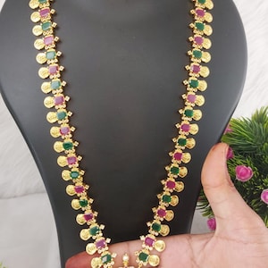 May include: A gold necklace with a pendant featuring a deity, surrounded by green and pink stones. The necklace is made of gold coins and has a matching pair of earrings.
