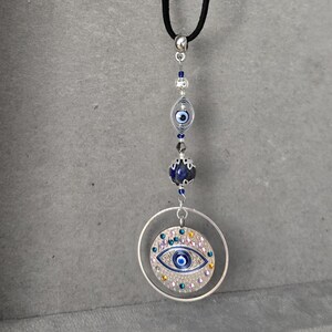 Op de afbeelding: Een zwarte koordketting met een zilverkleurige bedel met een blauw oogontwerp. De bedel is versierd met blauwe en iriserende kristallen. De ketting heeft ook een blauwe en een zilveren kraal.