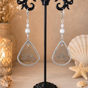 Peut inclure: Une paire de boucles d'oreilles pendantes argentées avec un cadre en forme de goutte d'eau présentant un motif découpé de coquillage ouvert contenant une perle. Chaque boucle est suspendue à un crochet avec une petite perle blanche et une perle plus grosse.