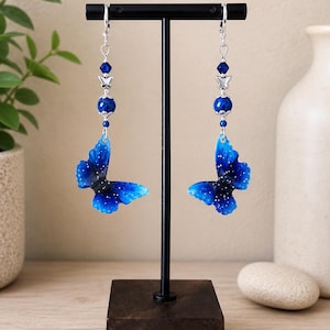 Op de afbeelding: Een paar bungelende oorbellen met blauwe vlindervormige bedels met witte spikkels. Elke oorbel bevat een zilverkleurig metalen vlinderaccent, een ronde blauwe kraal en een kleine gefacetteerde blauwe kristallen kraal aan een zilveren klapoorbel.