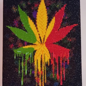 Puede incluir: Una pintura de diamantes de una hoja de cannabis en los colores verde, amarillo y rojo. La hoja tiene un fondo negro con motas coloridas. Los colores de la hoja gotean por el lienzo.