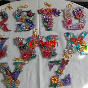 May include: Colorful beaded letter keychains. Each letter is adorned with beads in various colors, including red, blue, green, and yellow, and features floral and butterfly designs. The keychains have gold-colored clasps.