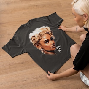 May include: Dark gray t-shirt featuring a portrait of a woman with blonde hair and a signature. The portrait is in shades of brown, beige, and white. The t-shirt is displayed on a wooden surface.
