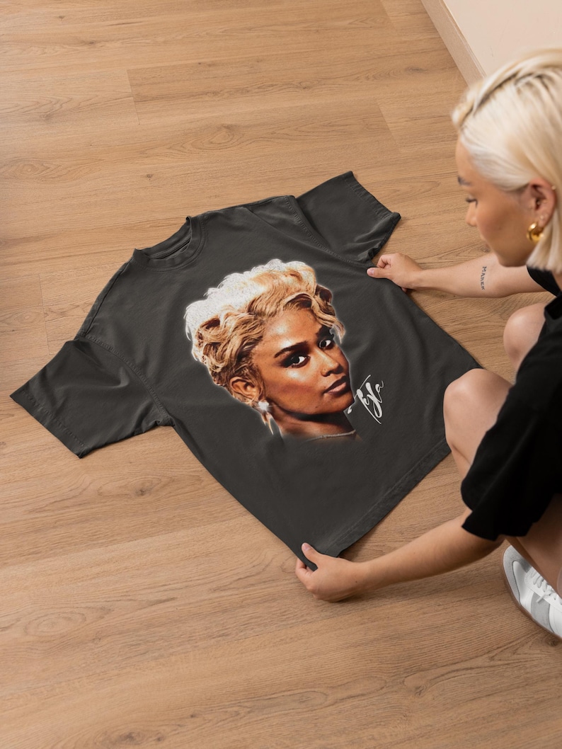 May include: Dark gray t-shirt featuring a portrait of a woman with blonde hair and a signature. The portrait is in shades of brown, beige, and white. The t-shirt is displayed on a wooden surface.