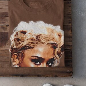 May include: A folded brown t-shirt featuring a close-up of a woman's face with blonde, curly hair and dark eyes. The shirt is on a wooden surface, with white sneakers visible in the foreground.