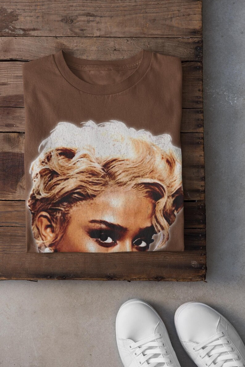 May include: A folded brown t-shirt featuring a close-up of a woman's face with blonde, curly hair and dark eyes. The shirt is on a wooden surface, with white sneakers visible in the foreground.