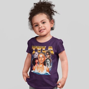 May include: A purple short-sleeved t-shirt with a vintage-style graphic of a woman's face with the text "TYLA" in gold letters.