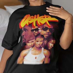 May include: Black t-shirt featuring a graphic print of a group of men. The graphic includes the word "USHER" in large, stylised letters. The men are wearing sunglasses and have a retro look.