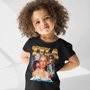 May include: A black t-shirt with a retro-style graphic featuring a collage of images of a young woman with the word "TYLA" in gold letters above her.