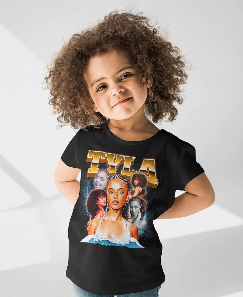 May include: A black t-shirt with a retro-style graphic featuring a collage of images of a young woman with the word "TYLA" in gold letters above her.