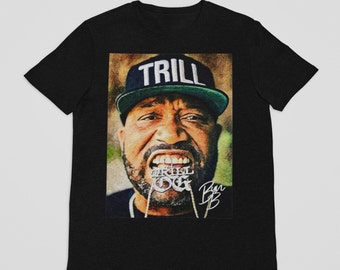 May include: Black t-shirt featuring a portrait of a man wearing a hat with the word "TRILL" and a silver chain with the words "TRILL OG". The image has a vintage, artistic style.