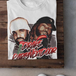 May include: White t-shirt with a graphic print of Drake and 21 Savage. The text "Drake x 21 Savage" is printed in red and white.