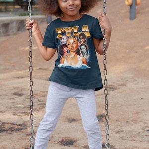 May include: A young girl wearing a navy blue t-shirt with a colorful graphic print of a woman's face and the word "TYLA" in gold lettering. She is sitting on a swing set.