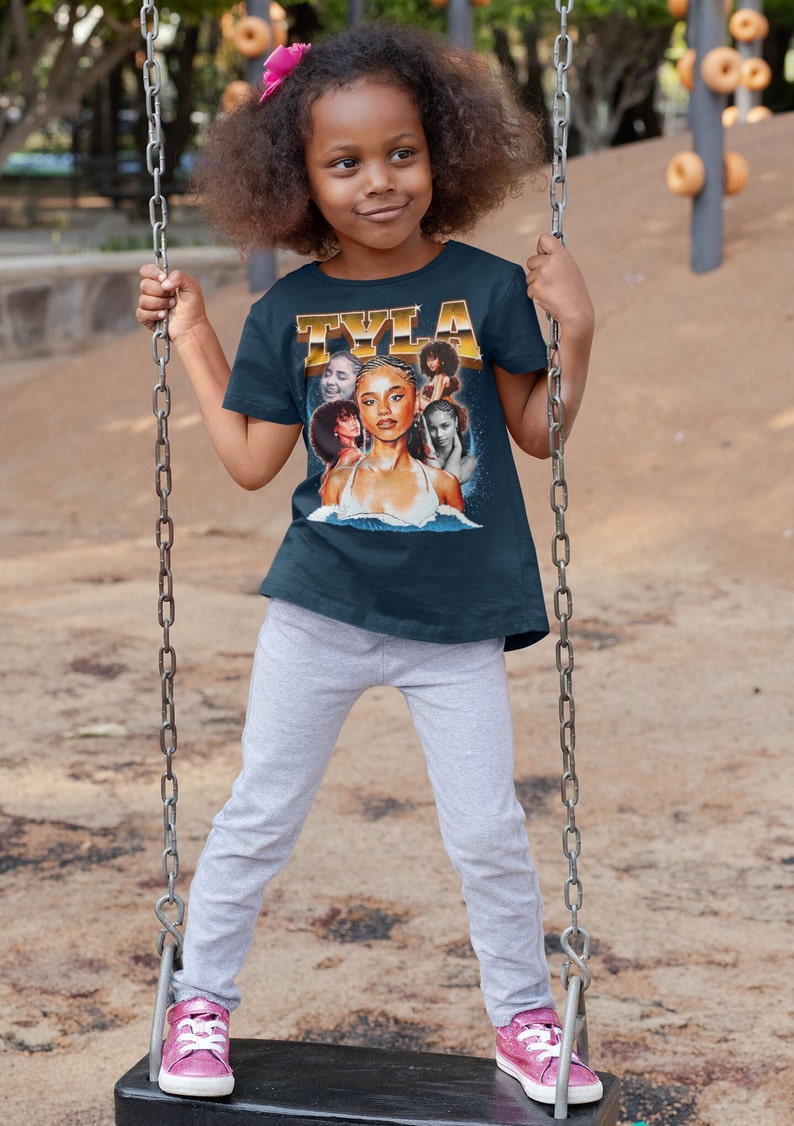 May include: A young girl wearing a navy blue t-shirt with a colorful graphic print of a woman's face and the word "TYLA" in gold lettering. She is sitting on a swing set.