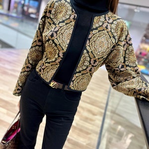 Könnte beinhalten: Eine schwarze, kurze Jacke mit einem goldenen und braunen geometrischen Muster. Die Jacke hat einen Stehkragen und lange Ärmel. Die Jacke wird über einem schwarzen Rollkragenpullover und einer schwarzen Jeans getragen.