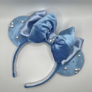 May include: A light blue headband featuring two round, plush ears adorned with faux pearls. A large, light blue satin bow is positioned between the ears. The headband itself is light blue.
