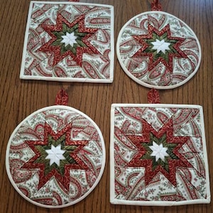 May include: Four quilted potholders featuring a central star pattern in white, dark green, and red, surrounded by a paisley print fabric in shades of red, green, and cream. Two are square and two are circular, each with a small fabric hanging loop.