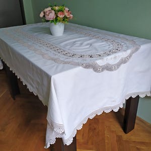 May include: A white rectangular tablecloth with a decorative lace border and a central oval design. A vase of pink and peach flowers sits on the table. The table has dark brown legs.