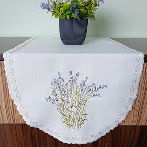 May include: White table runner with a lavender embroidery design. The runner has a scalloped edge with a decorative stitch. A dark gray pot with a lavender plant sits on the table.