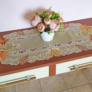 May include: An oval-shaped beige table runner with embroidered leaf designs in shades of orange, yellow, and brown. A white vase holds a bouquet of pink flowers, placed on a wooden table with white drawers.