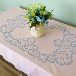 May include: A light pink tablecloth with a blue floral embroidered border. A small vase with blue and yellow flowers sits on the table. The tablecloth is rectangular and has a delicate, vintage aesthetic.