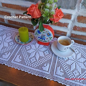 Può includere: Un runner da tavolo in crochet bianco con un motivo floreale. Il runner è su un tavolo di legno con un vaso di fiori, una candela e una tazza di caffè.