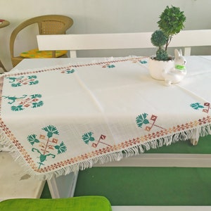 Peut inclure: Nappe blanche à bordure frangée et motifs floraux brodés en vert et marron. La nappe est posée sur une table blanche avec une plante verte dans un pot blanc et une figurine de lapin blanc.