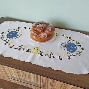 Peut inclure: Un chemin de table ovale blanc avec des motifs floraux brodés bleus et jaunes. Un pain sucré est sur une assiette blanche au centre du chemin de table.