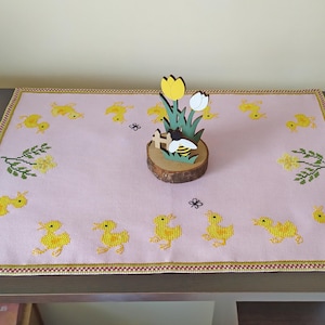 Op de afbeelding: Een roze tafelkleed met een gele geborduurde rand heeft een houten uitsnijding van een bij en tulpen. Het tafelkleed is versierd met geborduurde gele kuikens.