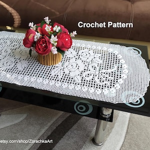 May include: White crochet table runner with a delicate floral pattern. The runner is on a black glass table with a vase of red and white flowers in the centre. The text "Crochet Pattern" is visible in the top right corner of the image.