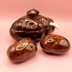 Può includere: Un set di saliera e pepiera in ceramica marrone con una zuccheriera abbinata con coperchio. Il set ha una finitura lucida e una forma organica unica. I salini e i pepini hanno dei piccoli fori sulla parte superiore.