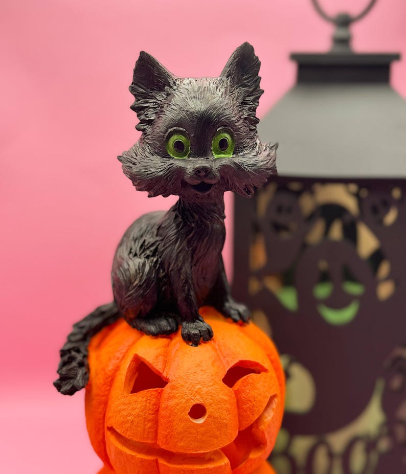 May include: A black cat figurine with bright green eyes sits atop an orange jack-o'-lantern. The cat has a fluffy appearance and is smiling. A black lantern is in the background, all against a pink backdrop. Halloween-themed decor.