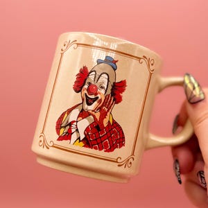 May include: A beige ceramic mug featuring a graphic illustration of a clown with a red nose, bright red hair, and a red plaid jacket. The clown is framed by a decorative brown border. The mug has a cylindrical body with a handle on the right side.