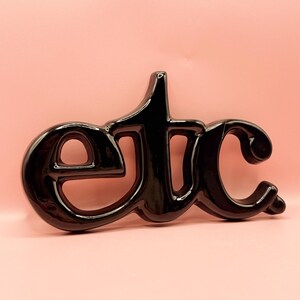 May include: A black, glossy decorative sign spelling "etc" in a cursive font. The letters are joined and have a smooth, reflective surface. The sign is set against a soft pink background.