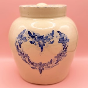 May include: Off-white ceramic jar with a lid, featuring a blue floral heart design. The jar has a rounded shape and a slightly textured surface. The lid has a small knob. The design is hand-painted with a signature at the bottom.