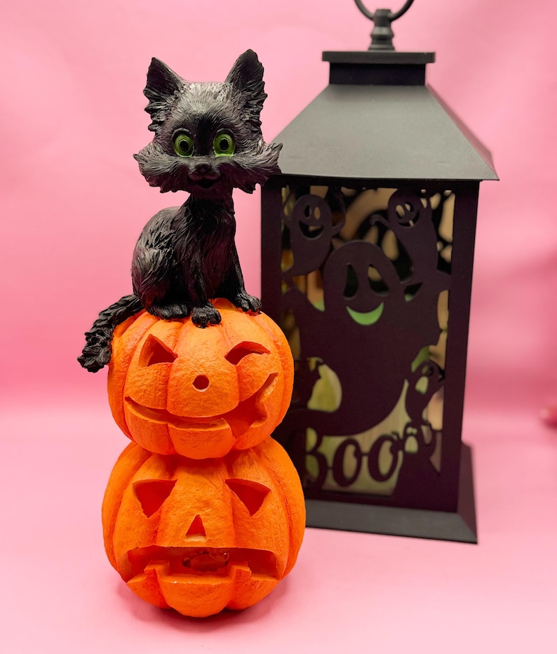 May include: A Halloween-themed decoration featuring a black cat figurine with green eyes perched atop two carved orange pumpkins. A black lantern with ghost cutouts and the word "BOO" is in the background. The pumpkins have carved faces.