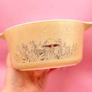 Puede incluir: Fuente para horno beige con borde y asas blancos. El plato presenta una ilustración marrón de setas, plantas y hierba. El plato se sostiene sobre un fondo rosa.