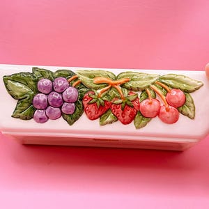 Puede incluir: Un plato rectangular de cerámica blanquecina con un diseño de frutas en relieve. El diseño incluye un racimo de uvas moradas, fresas rojas y cerezas rosas con hojas verdes. El plato es probablemente para servir o exhibir.
