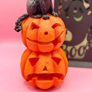 May include: A stacked orange Halloween decoration featuring two carved pumpkins with smiling faces. A black cat figurine sits atop the pumpkins. The background is a solid pink color.