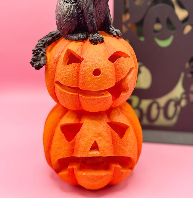 May include: A stacked orange Halloween decoration featuring two carved pumpkins with smiling faces. A black cat figurine sits atop the pumpkins. The background is a solid pink color.
