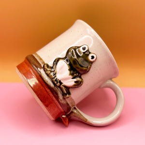 Könnte beinhalten: Eine Keramiktasse mit einem skurrilen Froschdesign. Die Tasse ist cremefarben mit einem braunen und roten Boden und Henkel. Ein dreidimensionaler Frosch mit grünen und weißen Details ist an der Seite der Tasse befestigt.
