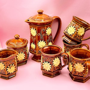 Puede incluir: Un juego de té de cerámica marrón con un diseño floral. El juego incluye una tetera, un azucarero, una crema y varias tazas. Las tazas están apiladas. El diseño floral es amarillo y crema.