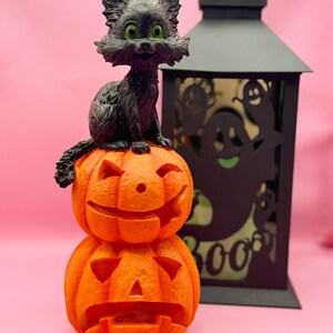 May include: A Halloween decoration featuring a black cat figurine with green eyes perched atop two stacked orange jack-o'-lanterns. A black lantern with ghost cutouts is in the background. The background is a solid pink color.